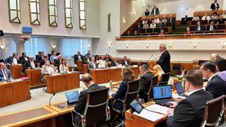 Ein Konferenzraum mit Menschen an Tischen, ein Mann steht am Podium und spricht, und mehrere Laptops und Papiere liegen auf den Tischen.