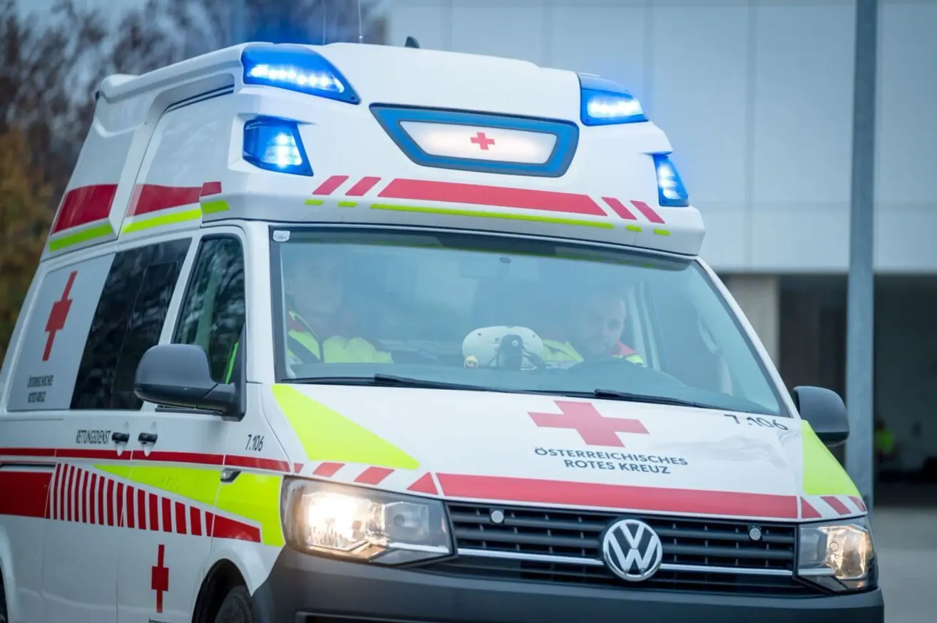 Ein Krankenwagenfahrzeug mit blauen Lichtern auf dem Dach und roten Lichtern vorne und hinten, mit dem Text 'Osterreichisches Rotes Kreuz' auf der Front. Zwei Personen sitzen im Inneren, einer fährt und der andere sitzt auf dem Beifahrersitz.