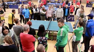 Eine Gruppe von Menschen ist um einen Tischtennistisch versammelt. Ein Mann spielt Tischtennis mit einer Frau in einer grünen Kapuzenjacke. Andere Leute schauen zu.