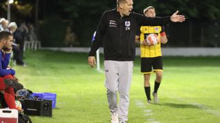 Ein Trainer in einer schwarzen Jacke mit Logo vorne dirigiert einen Spieler, der einen Fußball hält. Der Spieler trägt ein gelb-schwarzes Trikot. Sie sind auf einem Rasenfeld.