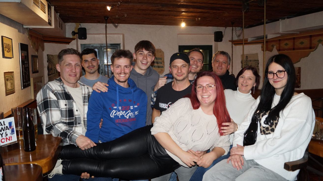 Eine Gruppe junger Erwachsener posiert für ein Foto in einem Raum mit Holzdecke, Lautsprechern und einer weißen Wand. Sie lächeln und stehen eng beieinander.