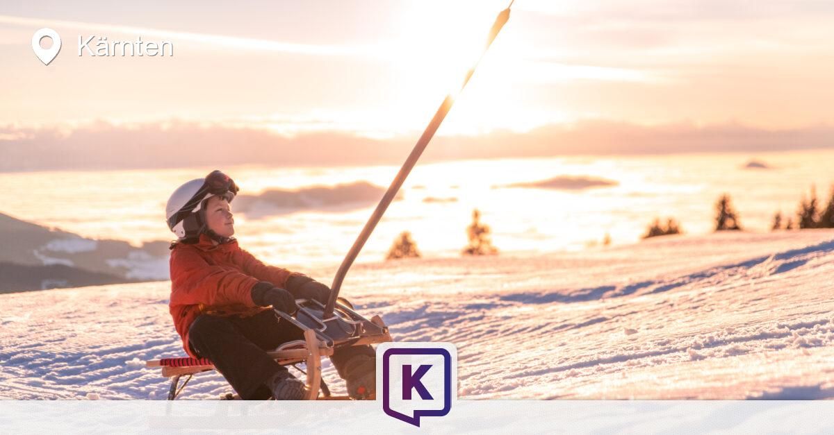 Eine Person sitzt auf einem Schlitten und hält einen langen Stab, trägt einen Helm und Handschuhe auf einem verschneiten Berg.