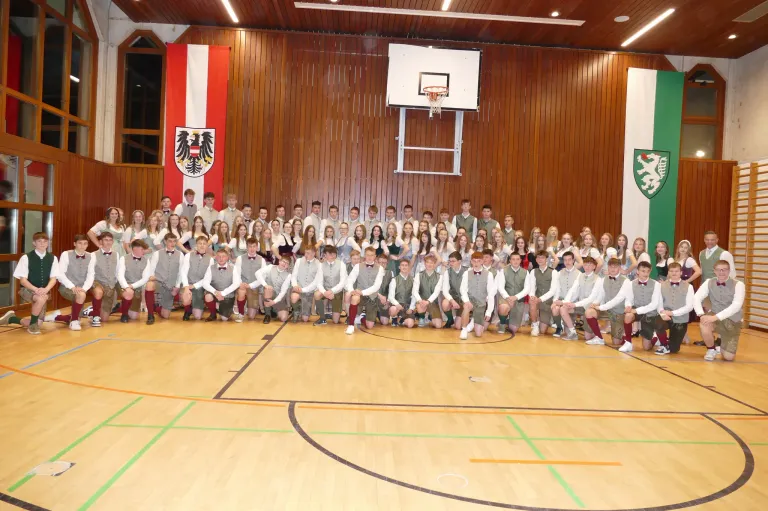 Eine große Gruppe junger Menschen in traditioneller österreichischer Tracht posiert für ein Foto in einer Turnhalle mit einem Basketballkorb und Fahnen im Hintergrund.