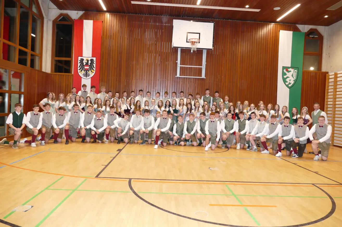 Eine große Gruppe junger Menschen in traditioneller österreichischer Tracht posiert für ein Foto in einer Turnhalle mit einem Basketballkorb und Fahnen im Hintergrund.