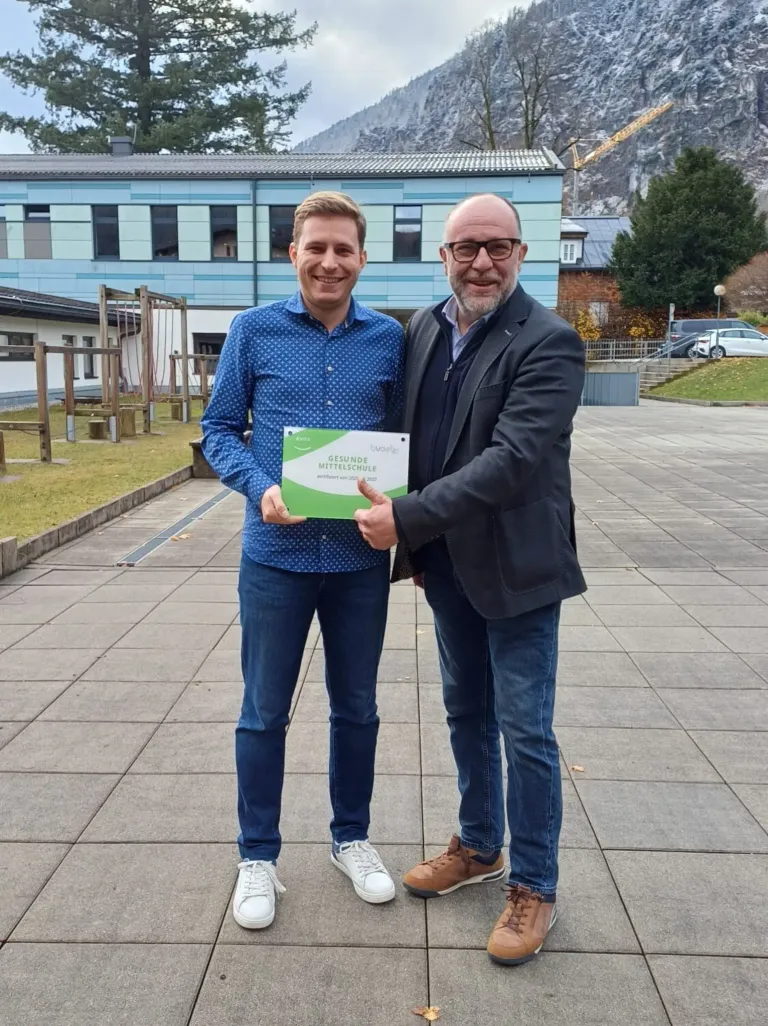 Zwei Männer stehen auf einer Betonplattform, einer präsentiert ein Zertifikat. Der Mann mit Brille hält das Zertifikat mit der rechten Hand. Hinter ihnen steht ein Gebäude mit einem Spielplatz.