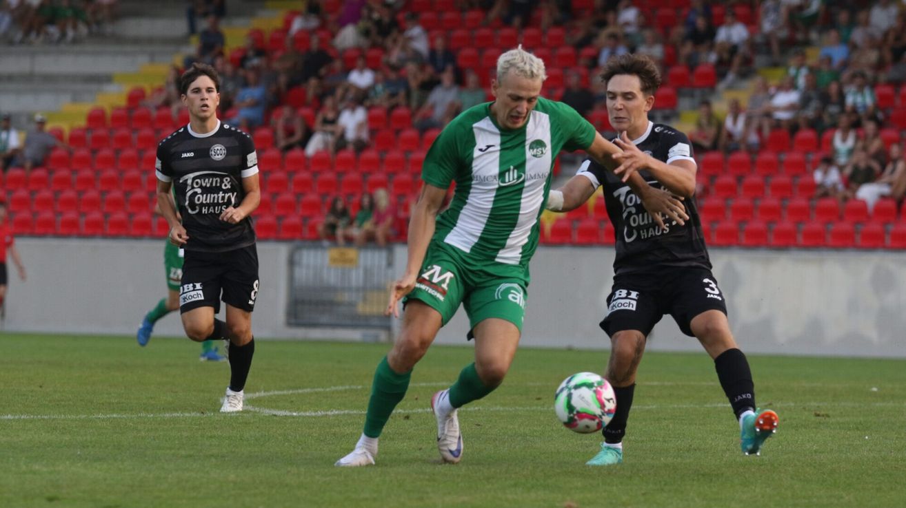 Drei Fußballspieler sind auf dem Spielfeld. Zwei kämpfen um den Ball, während der Dritte links steht. Das Feld ist von roten Tribünen umgeben, die mit Zuschauern gefüllt sind.
