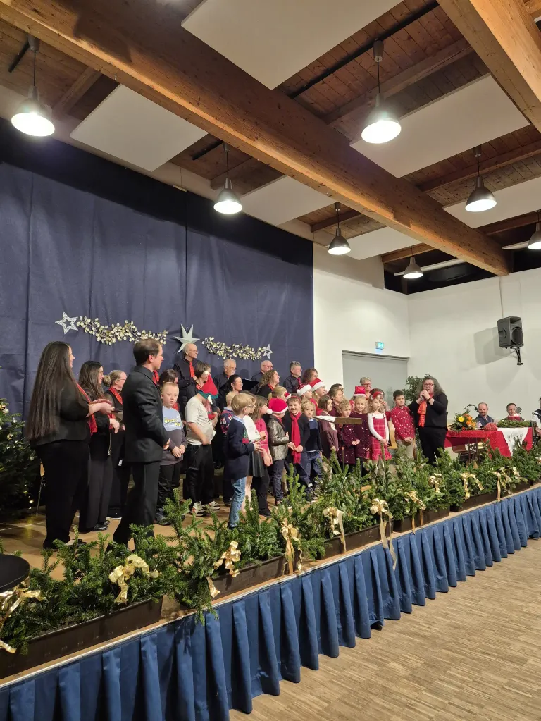 Ein Chor aus Kindern und Erwachsenen, die Weihnachtsmützen tragen, steht auf einer mit immergrünen Pflanzen geschmückten Bühne. Eine Frau leitet den Chor, während ein Mann neben ihnen steht. Im Hintergrund steht ein Weihnachtsbaum, und weitere Menschen sitzen auf Stühlen.
