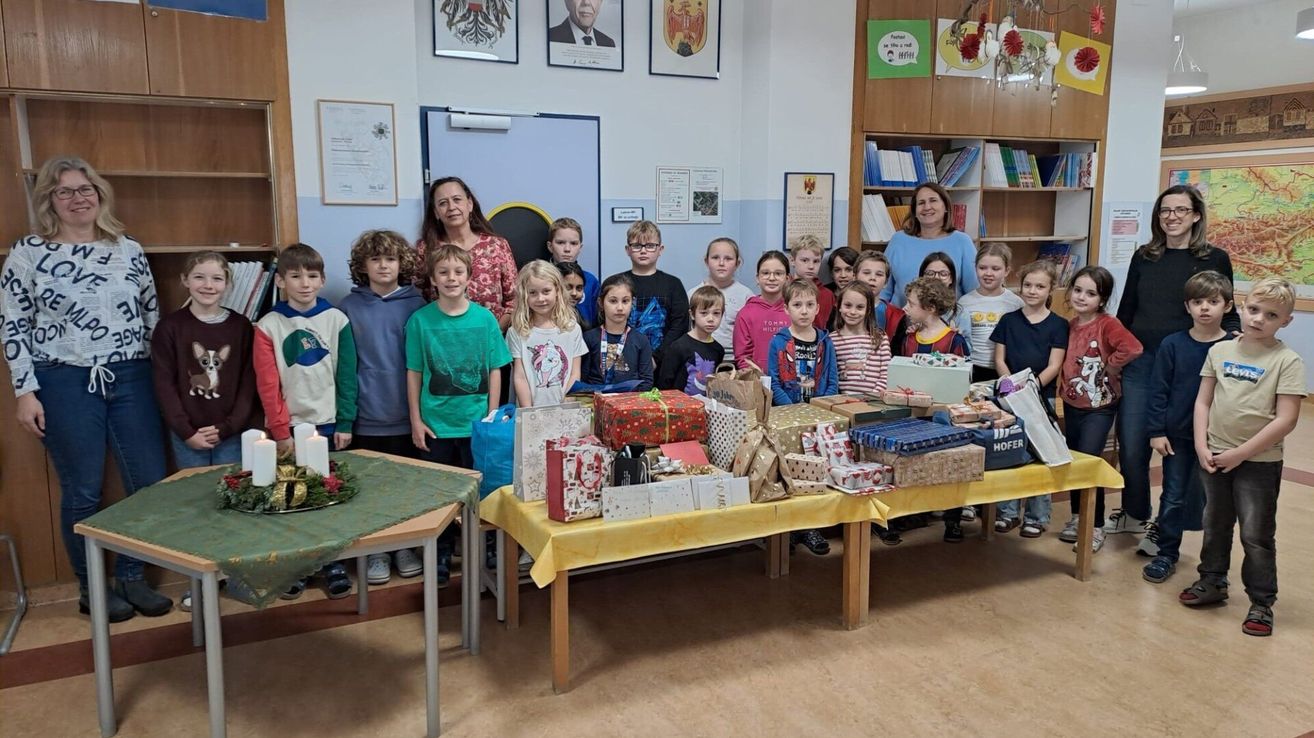 Eine Gruppe von Kindern und zwei Lehrern steht hinter einem Tisch voller verpackter Geschenke. Die Kinder lächeln und posieren für ein Foto.