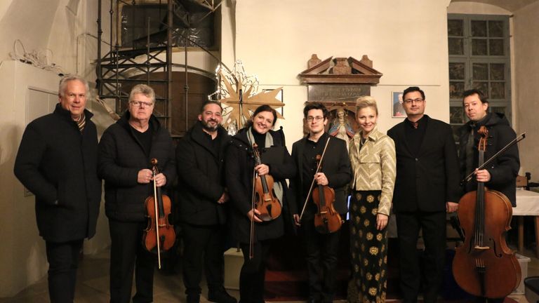 Eine Gruppe von Musikern mit Violinen posiert für ein Foto vor einer Kircheninnenansicht. Sie lächeln und scheinen bereit für eine Aufführung zu sein.