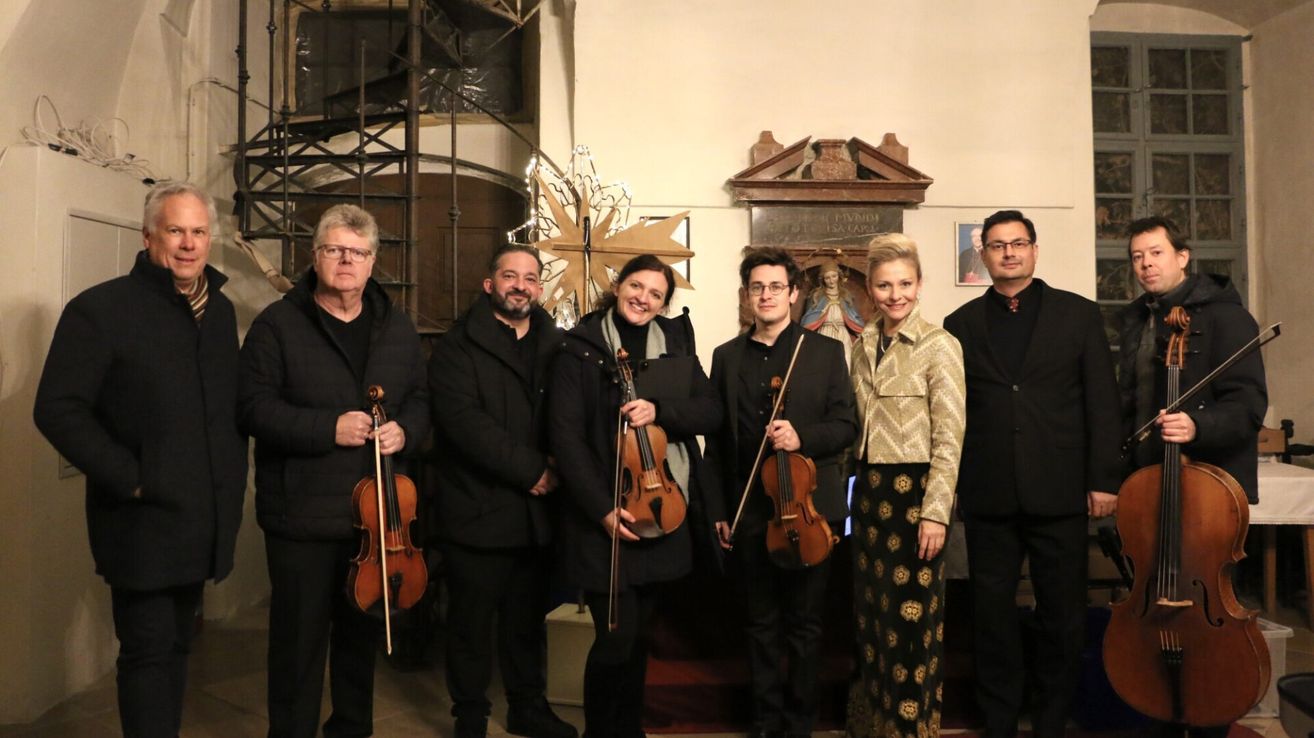 Eine Gruppe von Musikern mit Violinen posiert für ein Foto vor einer Kircheninnenansicht. Sie lächeln und scheinen bereit für eine Aufführung zu sein.