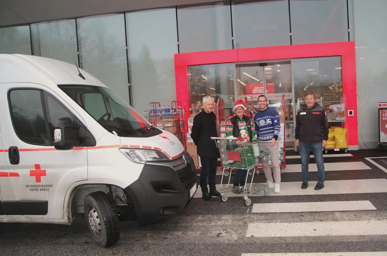 Vier Personen stehen vor einem Geschäft. Zwei Männer vorne halten einen Einkaufswagen. Ein Lieferwagen ist links geparkt.