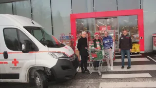 Vier Personen stehen vor einem Geschäft. Zwei Männer vorne halten einen Einkaufswagen. Ein Lieferwagen ist links geparkt.