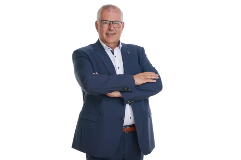 Ein Mann in einem dunkelblauen Anzug und mit einer Brille steht mit verschränkten Armen und lächelt.