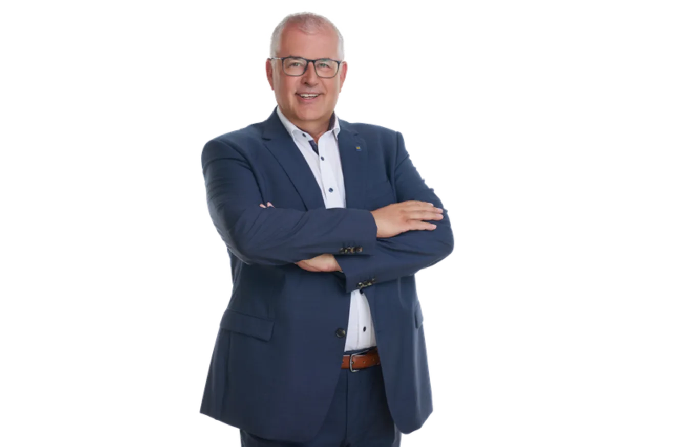 Ein Mann in einem dunkelblauen Anzug und mit einer Brille steht mit verschränkten Armen und lächelt.