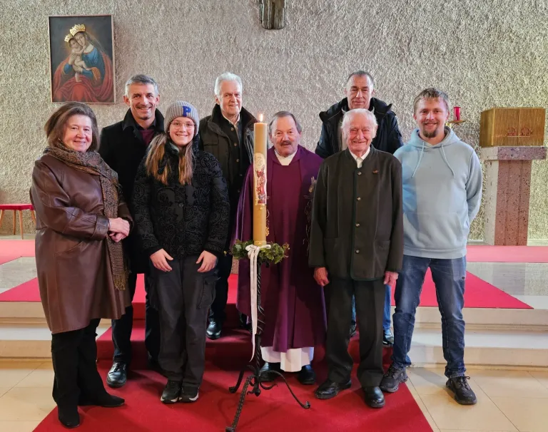 Eine Gruppe von Menschen ist in einer Kirche versammelt und posiert für ein Foto. Ein Priester steht neben einer brennenden Kerze auf einem Ständer. Ein religiöses Bildnis ist an der Wand hinter ihnen.