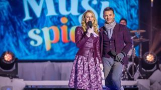 Eine Frau in einem lila Kleid und ein Mann in einem lila Anzug stehen auf der Bühne, lächeln und halten Mikrofone. Hinter ihnen befindet sich ein großes Banner mit dem Wort 'Spider'.