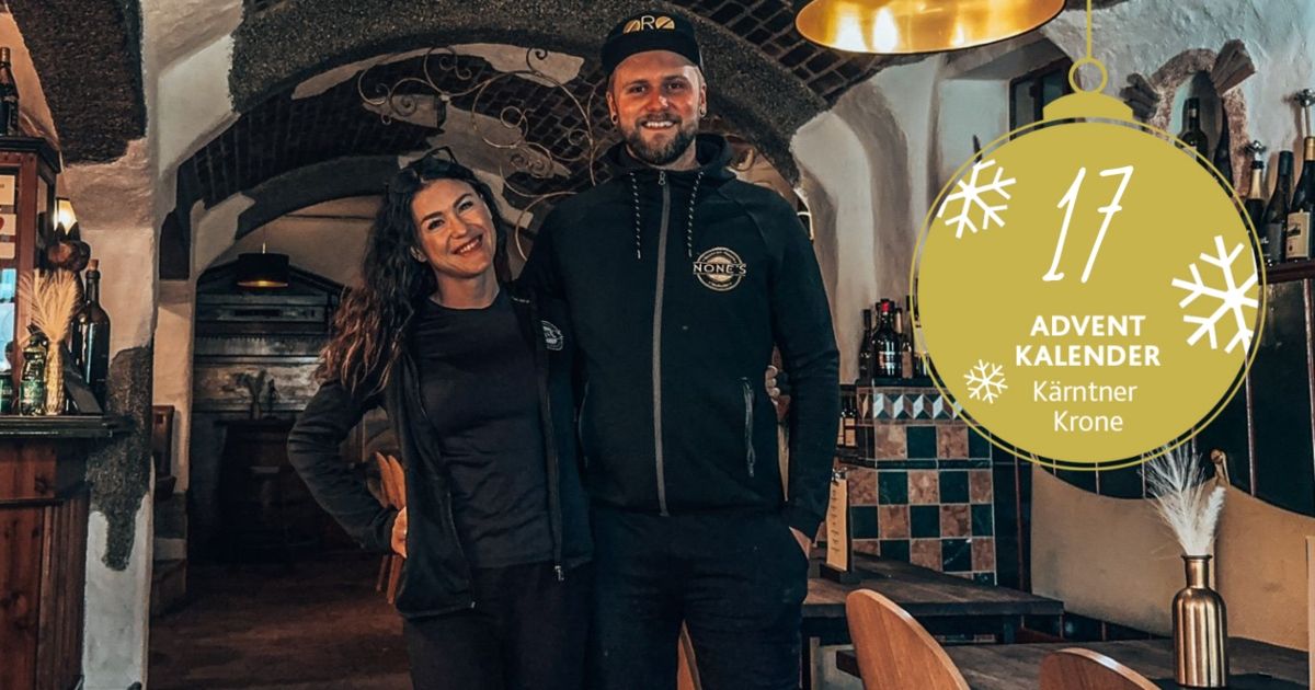 Ein Mann und eine Frau stehen lächelnd in einem Restaurant mit gefliester Wand. Hinter ihnen befindet sich ein Regal mit Flaschen. An der Decke hängt eine Lampe.