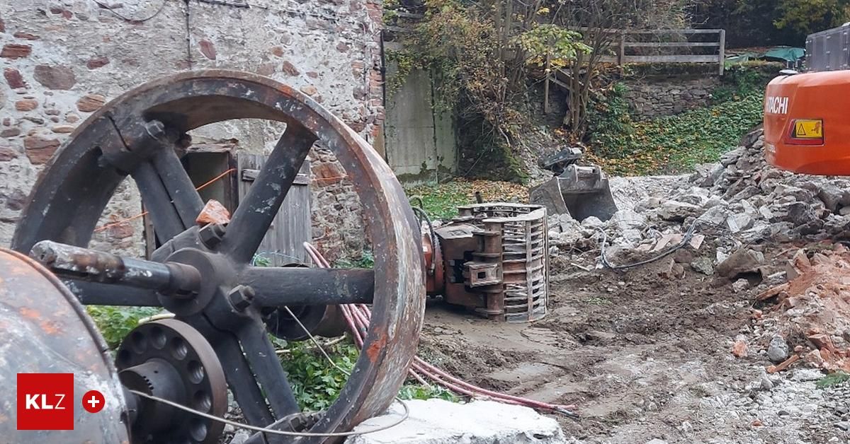 Eine Außenszene zeigt ein großes, rostiges Rad neben einem Schutthaufen. Ein Baufahrzeug ist in der Nähe geparkt. Hinter dem Schutt befindet sich eine Steinmauer mit viel Grün.