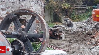 Eine Außenszene zeigt ein großes, rostiges Rad neben einem Schutthaufen. Ein Baufahrzeug ist in der Nähe geparkt. Hinter dem Schutt befindet sich eine Steinmauer mit viel Grün.