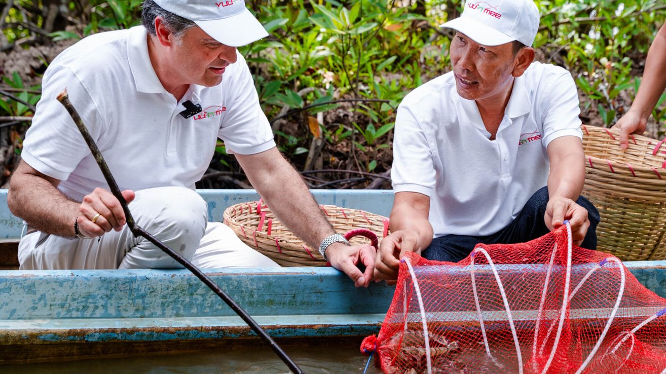 Zwei Männer in weißen Hemden und Hüten sitzen in einem blauen Boot und halten ein rotes Netz mit Fischen. Das Boot befindet sich in einem Fluss mit grünen Pflanzen im Hintergrund.