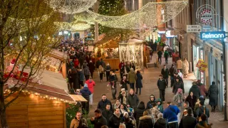 Eine überfüllte Straße voller Menschen, geschmückt mit festlichen Lichtern und Weihnachtsständen. Es ist Nacht, und Personen gehen oder stehen, einige tragen Kinder.