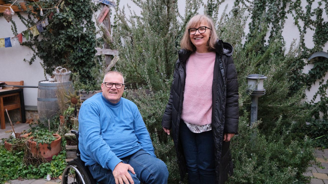 Ein Mann im Rollstuhl und eine Frau mit Brille und Jacke lächeln in einem Garten mit Sträuchern.