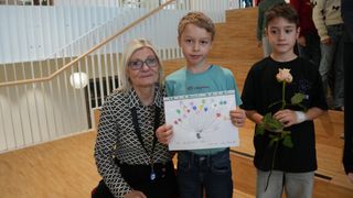 Eine ältere Frau mit Brille steht neben einem jungen Jungen, der ein Papier mit einer farbenfrohen Zeichnung in der Hand hält. Sie lächeln beide. Hinter ihnen steht ein weiterer Junge, der eine Pflanze in der Hand hält. Im Hintergrund befindet sich eine Treppe.