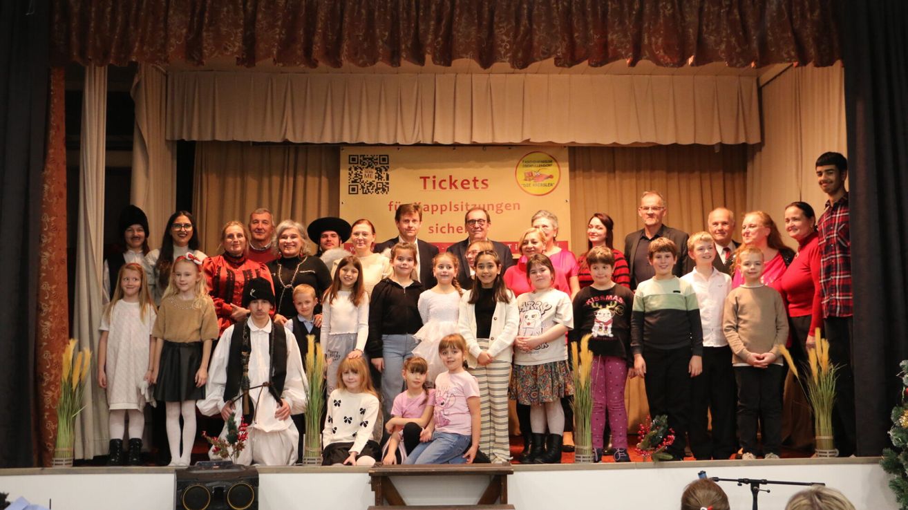Eine Gruppe von Erwachsenen und Kindern posiert für ein Foto auf der Bühne mit einem Banner, das 'Tickets' und einen QR-Code zeigt. Die Kinder sind in verschiedenen Kostümen gekleidet.