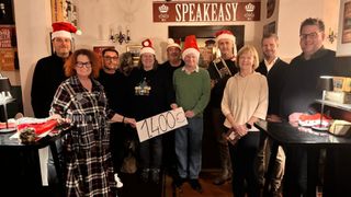 Eine Gruppe von Menschen posiert für ein Foto in einer Bar. Sie tragen Weihnachtsmützen. Eine Person hält ein Schild mit der Nummer 1400. Hinter ihnen ist eine Wand mit einem Schild, auf dem 'SPEAKEASY' steht.