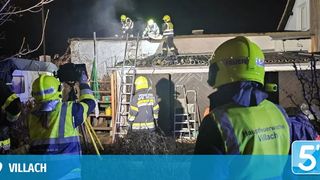 Feuerwehrleute in gelben und grünen Uniformen sind auf dem Dach eines Gebäudes mit Rauch, in der Nacht. Einer ist auf einer Leiter und andere sind am Boden. Eine Kamera wird von einer Person gehalten.