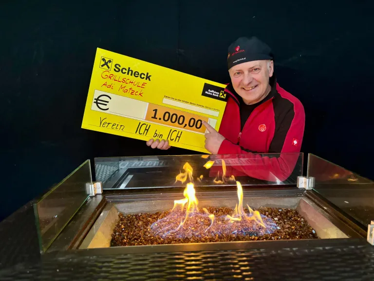 Ein Mann in einem roten Jacken hält einen gelben Scheck über 1.000 Euro und steht neben einem Kamin mit Flammen.