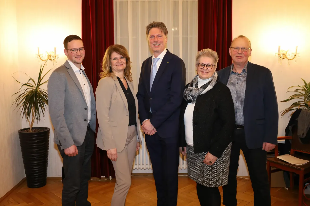 Fünf Personen stehen vor einem weißen Heizkörper mit roten Vorhängen im Hintergrund. Sie alle lächeln und scheinen für ein Foto zu posieren.