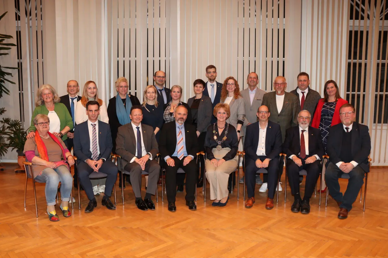 Eine Gruppe von Fachleuten in formeller Kleidung, einschließlich Anzüge und Krawatten, posiert für ein Foto auf einem Holzboden. Sie sitzen auf Stühlen und stehen vor einer Kulisse aus weißen Jalousien.