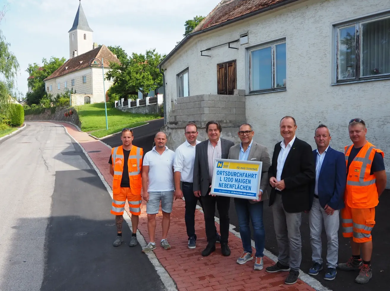 Eine Gruppe von Männern in Anzügen und mit Brillen steht vor einem Gebäude mit einem Schild, das 'Ortsdurchfahrt L 1200 Maigen Nebenflächen' besagt.