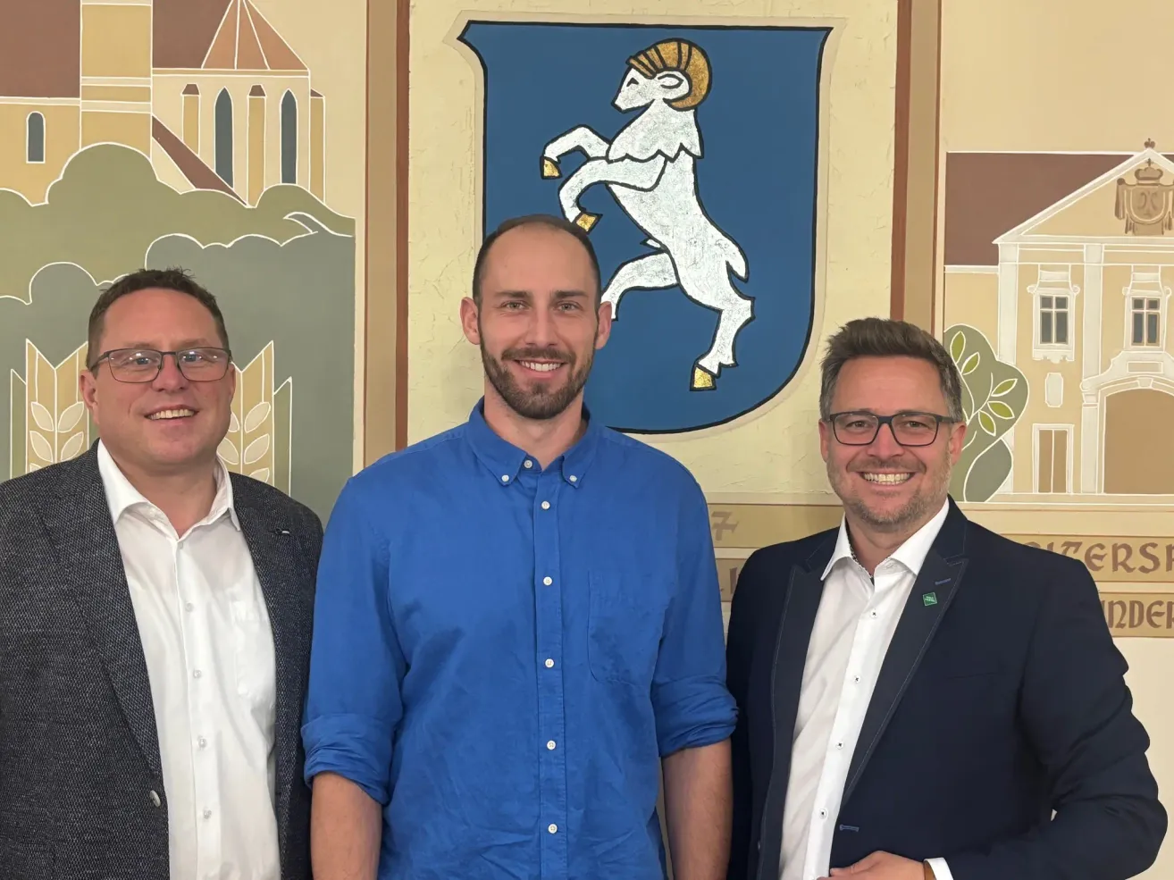 Drei Männer stehen zusammen vor einer Wandmalerei. Der mittlere Mann trägt ein blaues Hemd, die anderen tragen Anzüge. Ein Wappenschild steht hinter ihnen.
