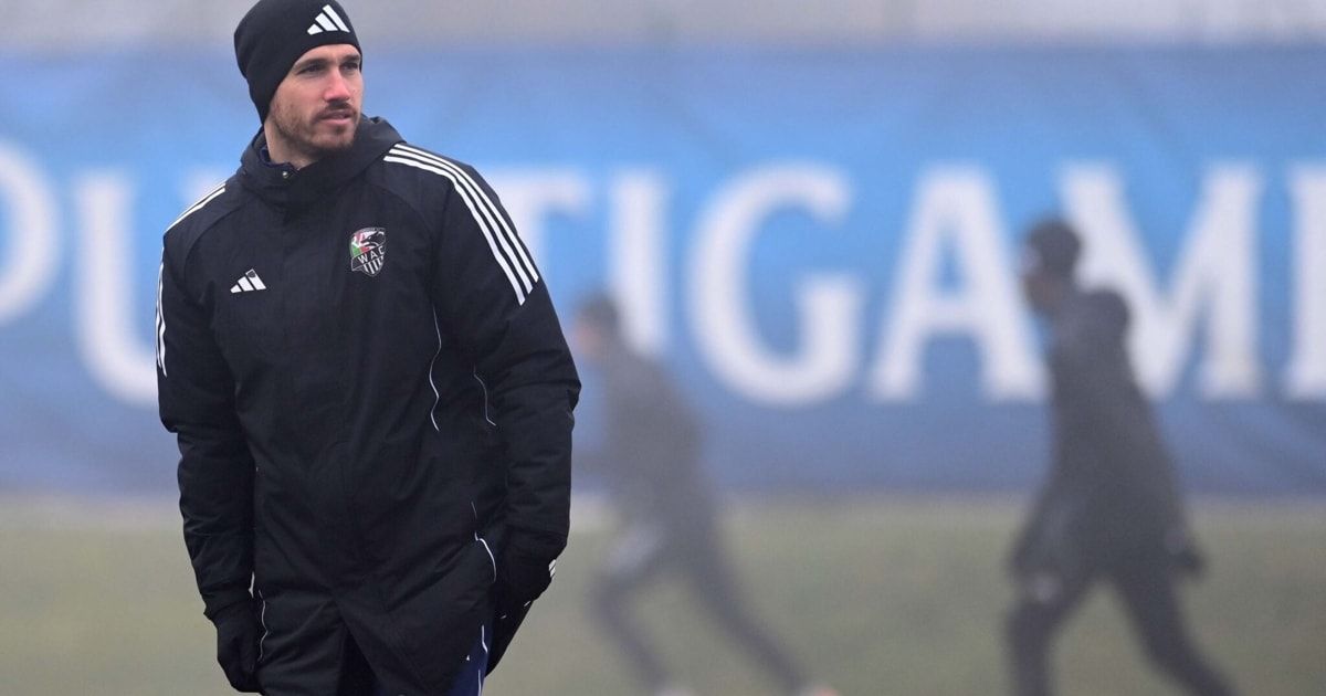 Ein Mann in einer schwarzen Adidas-Jacke mit drei Streifen an jedem Ärmel, einer schwarzen Mütze und einem unscharfen Hintergrund mit dem Wort 'GA'.