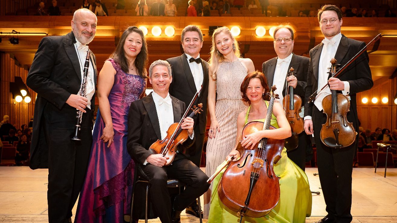 Eine Gruppe klassischer Musiker, darunter Geiger und ein Cellist, posiert für ein Foto in einem Konzertsaal. Sie tragen alle formelle Kleidung, und einige lächeln.