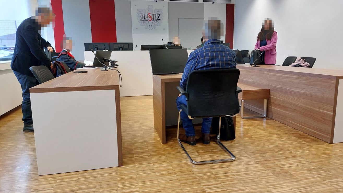 Eine Person sitzt mit verschwommenem Gesicht in einem Gerichtssaal an einem Schreibtisch mit Monitoren und Mikrofonen. Eine Frau steht im Hintergrund.