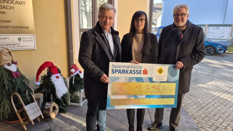 Drei Menschen stehen vor einem Gebäude und lächeln. Ein Mann hält einen großen Scheck über 1.395 €, mit zwei Frauen neben ihm. Dahinter sind festliche Dekorationen und ein Fenster.