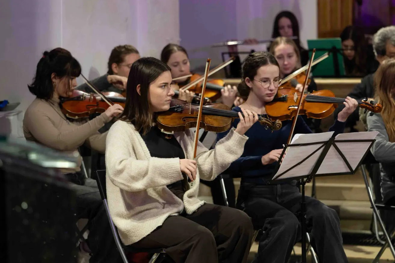 Eine Gruppe junger Frauen sitzt auf Stühlen und spielt Violinen bei einer Probe. Sie sind lässig gekleidet und haben Notenständer vor sich. Die Violinen sind braun, und die Frauen konzentrieren sich auf ihre Instrumente.