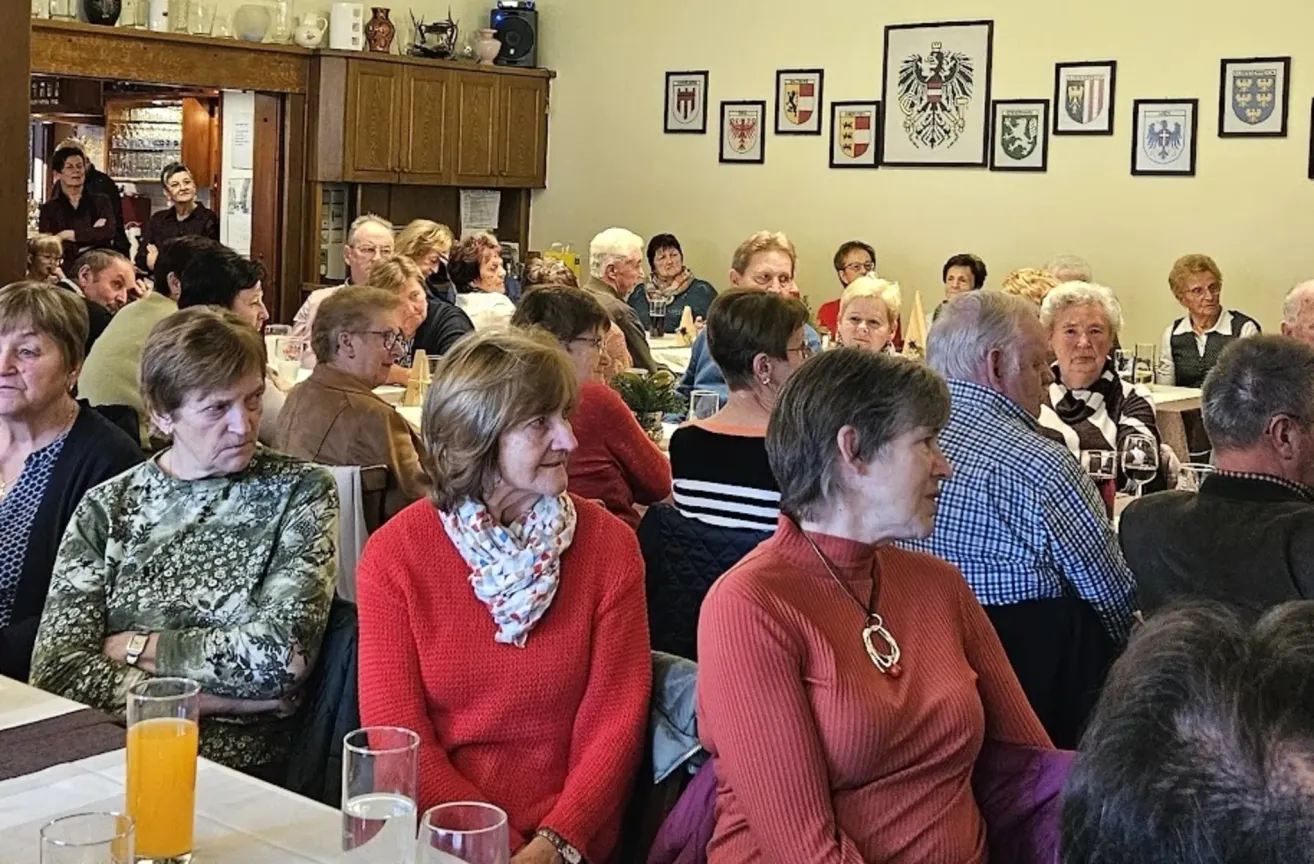 Eine Gruppe älterer Menschen sitzt in einem Speisesaal an einem Tisch, einige mit Brillen und Schals, mit dekorativen Rahmen und einem Schrank im Hintergrund.