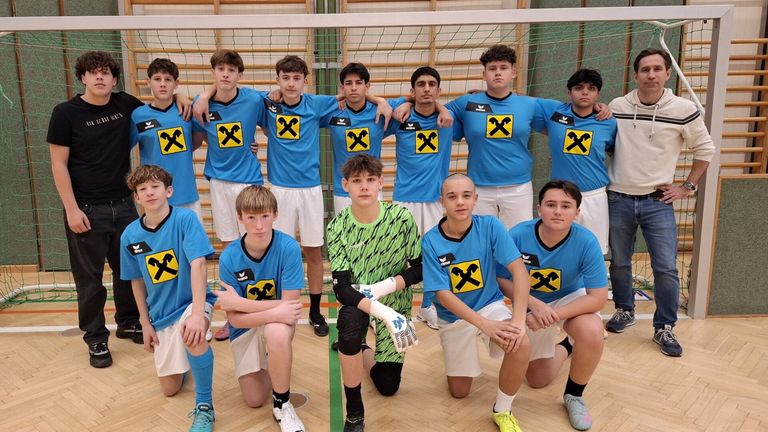 Eine Gruppe junger Männer in blauen und weißen Uniformen, möglicherweise ein Sportteam, posiert für ein Foto vor einem Netz. Sie stehen und hockten, einige von ihnen mit den Händen auf den Schultern. Der Boden ist aus Holz, und dahinter befindet sich eine Holzwand.