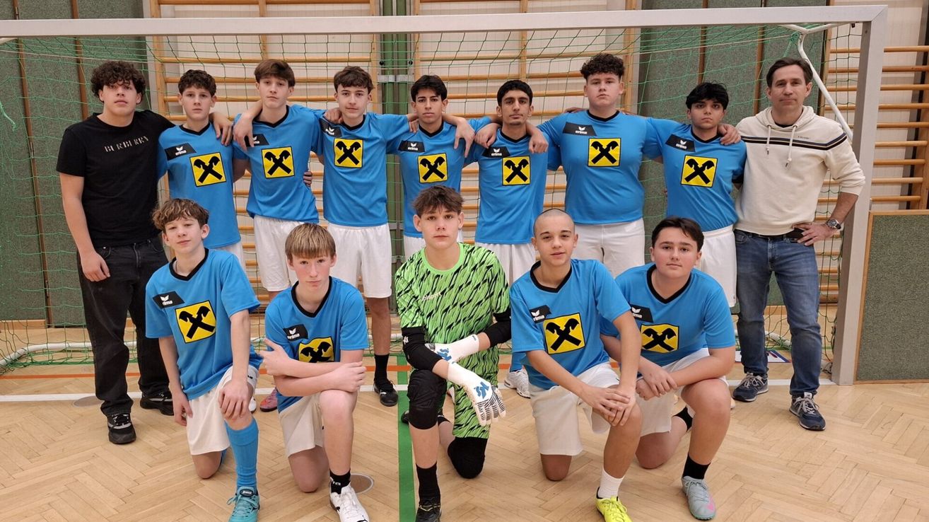 Eine Gruppe junger Männer in blauen und weißen Uniformen, möglicherweise ein Sportteam, posiert für ein Foto vor einem Netz. Sie stehen und hockten, einige von ihnen mit den Händen auf den Schultern. Der Boden ist aus Holz, und dahinter befindet sich eine Holzwand.