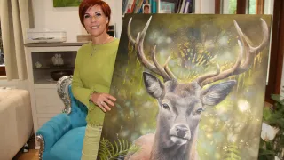 Eine Frau in einem grünen Oberteil steht neben einem großen Gemälde eines Hirsches. Das Gemälde zeigt detailliert die Hörner und ausdrucksstarken Augen des Hirsches. Hinter ihr befindet sich ein Raum mit Bücherregalen, einem Stuhl und einer kleinen Statue.