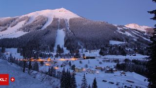Ein verschneiter Bergdorf mit Kiefern, Häusern und einer Skipiste. Der Himmel ist blau mit einigen Wolken.