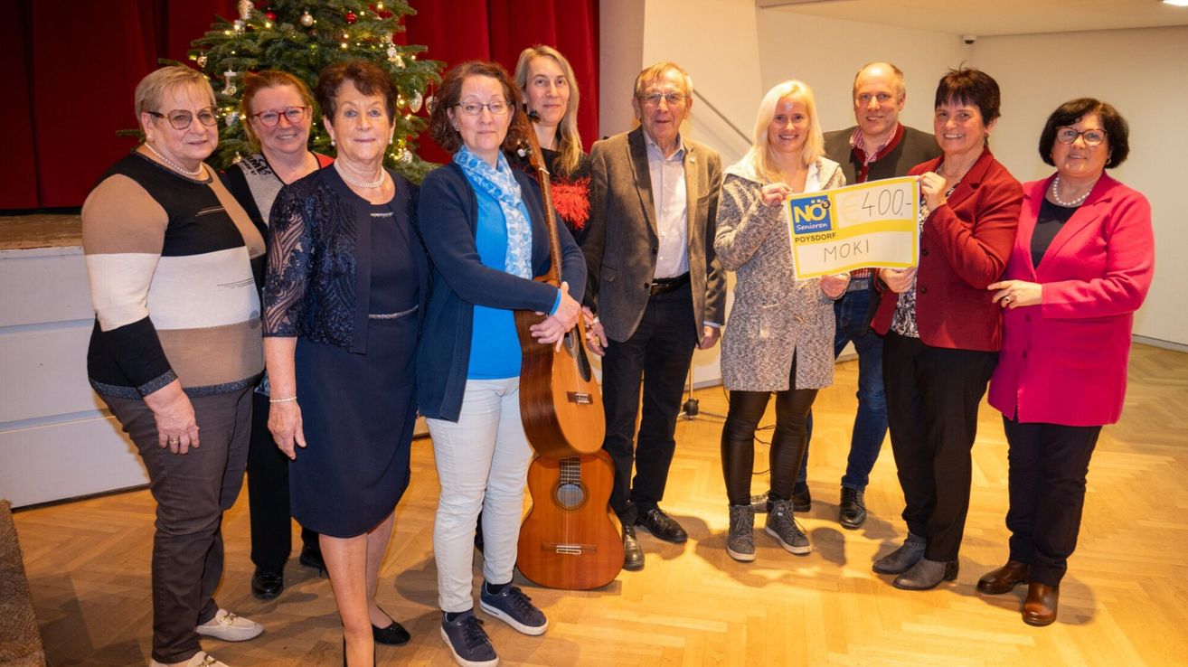 Eine Gruppe Erwachsener steht lächelnd zusammen im Innenbereich, einer hält ein Schild mit der Aufschrift 'NEIN 400- MOK1'. Ein Weihnachtsbaum und ein roter Vorhang sind im Hintergrund.