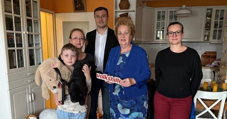 Eine Familie steht in einer Küche zusammen mit einem Schild, das Volkshilfe sagt. Ein Junge hält zwei Kuscheltiere.