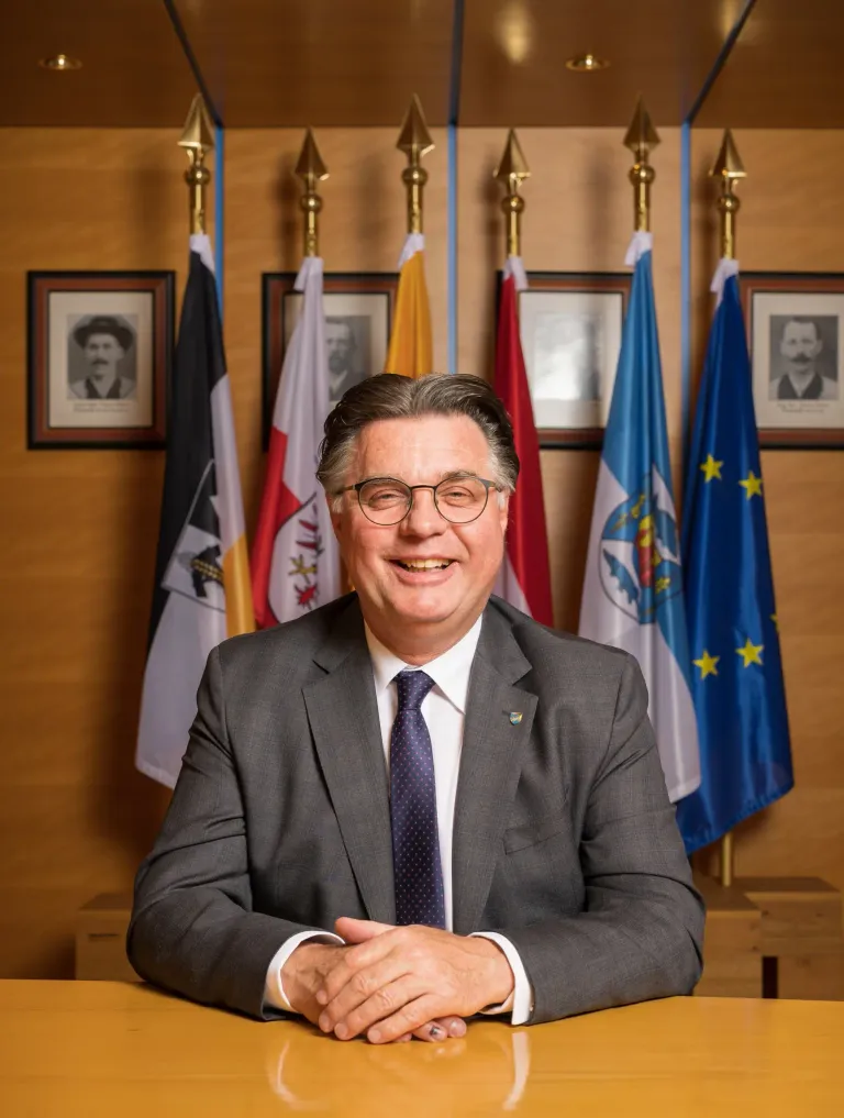 Ein Mann in Anzug und Krawatte lächelt und sitzt vor Flaggen und Porträts.