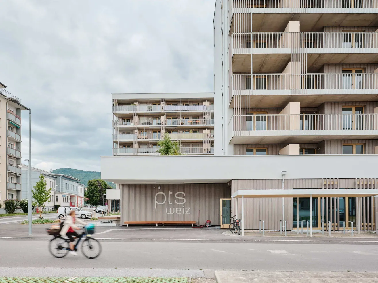 Eine Person fährt ein Fahrrad vor einem Gebäude mit einer Bank und dem Wort 'pts weiz'.