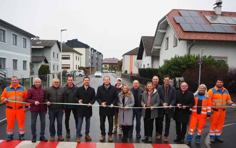 Eine Gruppe von Menschen steht auf einer Straße und hält ein Band, wahrscheinlich zur Feier eines Ereignisses. Dahinter befinden sich Häuser mit Solarpanelen auf den Dächern.